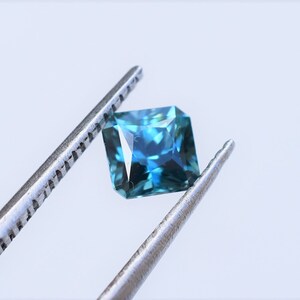 Op de afbeelding: Een vierkant geslepen edelsteen met een diep blauw en groenblauw kleurenpalet, voorzien van meerdere facetten die het licht reflecteren. De steen wordt vastgehouden door een zilverkleurige metalen pincet, wat de transparantie benadrukt.