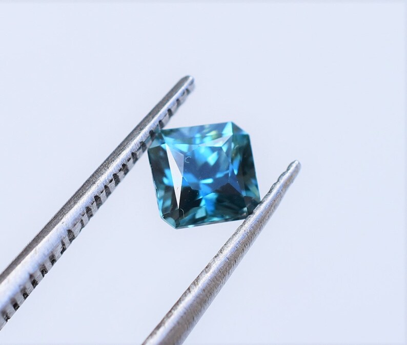Op de afbeelding: Een vierkant geslepen edelsteen met een diep blauw en groenblauw kleurenpalet, voorzien van meerdere facetten die het licht reflecteren. De steen wordt vastgehouden door een zilverkleurige metalen pincet, wat de transparantie benadrukt.