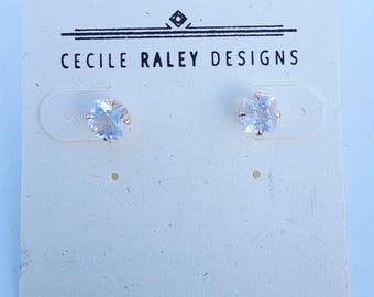 Puede incluir: Un par de pendientes de botón con gemas transparentes de corte cuadrado montadas en garras de metal, sobre una tarjeta de presentación blanca con el texto CECILE RALEY DESIGNS y un pequeño logotipo en forma de diamante en tinta negra.