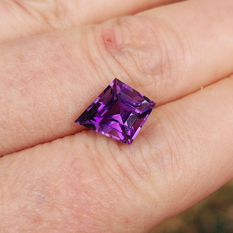 May include: A kite-shaped, faceted gemstone with a deep, vibrant purple color. The stone features a symmetrical geometric cut with sharp angles and multiple reflective surfaces that catch the light, displaying varying shades of violet and dark purple.
