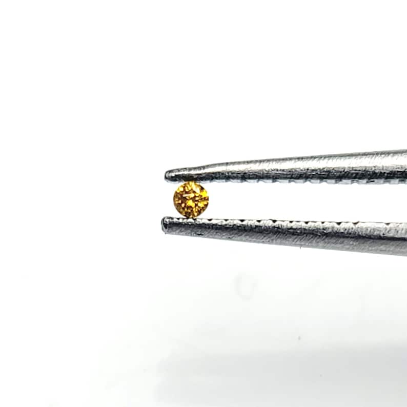 May include: A small, round, faceted gemstone with a vibrant amber-orange colour held securely between the serrated metal tips of a pair of silver-toned tweezers. The stone displays a brilliant, reflective surface with multiple cut facets.