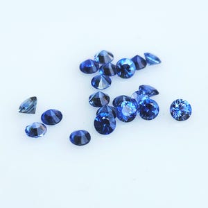 Puede incluir: Un conjunto de gemas azules brillantes dispersas sobre una superficie blanca. Las gemas varían en forma, algunas son redondas y otras facetadas. El color azul intenso de las piedras crea un contraste llamativo con el fondo claro.