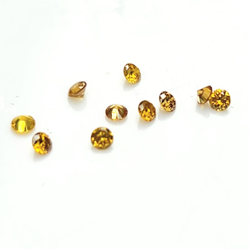 May include: A collection of ten small, round, faceted gemstones in a vibrant golden-yellow hue. Each stone features a brilliant cut with multiple reflective surfaces that catch the light, creating a sparkling appearance across the scattered arrangement.