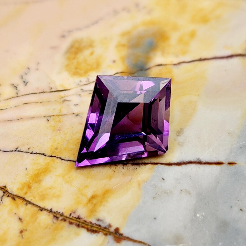 May include: A kite-shaped, faceted gemstone with a deep purple color. The stone features a geometric cut with multiple triangular and trapezoidal facets that reflect light, creating a sharp, angular silhouette with a pointed top and bottom.