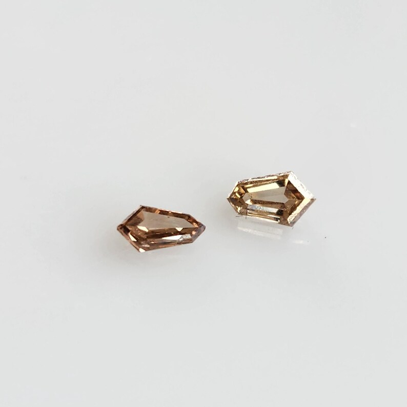 May include: Two loose shield-cut gemstones with a warm brown hue and faceted surfaces. These stones exhibit a translucent quality with sharp, geometric edges and a polished finish, reflecting light across their multiple angled planes.