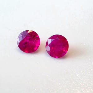 May include: Two round, faceted, deep red gemstones, likely rubies, are displayed against a white background. The gems have a brilliant, polished appearance, reflecting light with multiple facets. The stones are of similar size and shape.