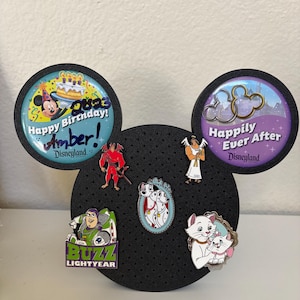 May include: A black Mickey Mouse-shaped pin display board with various Disney character pins. The pins include Buzz Lightyear, Marie and Berlioz, a Dalmatian pair, and others. Two large circular pins at the top read "Happy Birthday! Amber!" and "Happily Ever After."