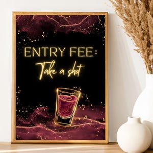 以下が含まれることがあります： 黒い背景に「ENTRY FEE: Take a shot」と金色の文字が書かれた額入りのアートプリント。画像には赤い液体が入ったショットグラスが描かれています。背景はバーガンディとゴールドの抽象的なデザインです。