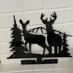 Może przedstawiać: Czarna metalowa grafika ścienna przedstawiająca sylwetki dwóch jeleni, drzew i gór. Jelenie stoją obok siebie. Dzieło sztuki ma poziomy pasek z dwoma otworami do montażu na białej ceglanej ścianie.