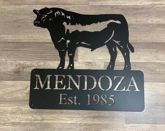 May include: Black metal bull silhouette wall art with the name "MENDOZA" and "Est. 1985" cut out on a rectangular sign. The bull is standing above the sign on a wooden background.