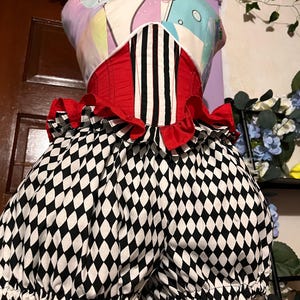 以下が含まれることがあります： パステルカラーのボディスと中央の黒と白のストライプパネルが特徴のコルセットトップとブルマーのセット。ウエストには赤いフリルのトリムが施され、ボリュームのあるブルマーには黒と白のダイヤモンド柄があしらわれています。