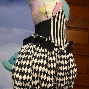 Op de afbeelding: Een zwart-witte harlekijnpatroonjurk met een gestreept lijfje en een zwarte top in korsetstijl. De jurk heeft een gerimpelde halslijn en een volle, geplooide rok. De jurk wordt getoond op een mannequin met een kleurrijke, bedrukte kop.