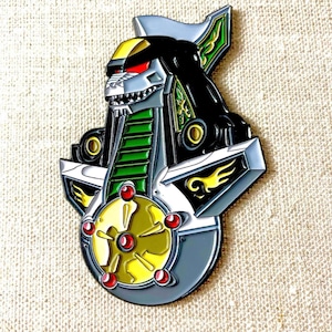 May include: A green, black, and silver enamel pin featuring a robot with a lion head. The robot has a large, round, gold-colored wheel with red accents.