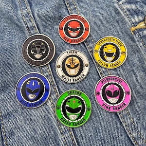 May include: A set of seven circular enamel pins featuring Power Rangers helmets in red, black, white, yellow, blue, green, and pink. Each pin displays the character name, the corresponding dinosaur or creature, and the ranger color in bold text.