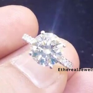 May include: A sparkling diamond ring with a large, round-cut center stone. The band is adorned with smaller accent stones. The ring is held up against a dark background. The text "EtherealJewelsCA" is visible.