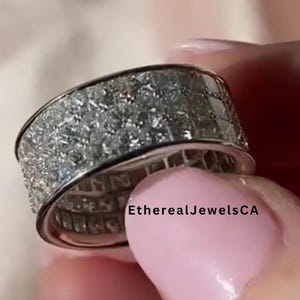 May include: A close-up of a silver ring with a band covered in small, square-cut, clear gemstones. The ring is held in a hand with pink nail polish. The text "EtherealJewelsCA" is visible.