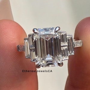 May include: A silver ring with an emerald-cut center stone and baguette side stones. The ring is held between two fingers. The text "EtherealJewelsCA" is visible on the ring.