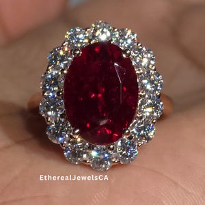 Peut inclure: Une bague en or ornée d'une grosse pierre rouge foncé de forme ovale, entourée de petits diamants transparents. La bague est présentée sur un fond neutre, mettant en valeur l'éclat des pierres. Le texte "EtherealJewelsCA" est visible.