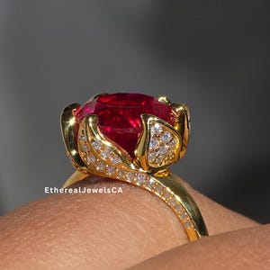 May include: A gold ring featuring a large, oval-cut, vibrant red gemstone. The ring's setting has a floral design with small, sparkling accent stones. The band is also adorned with smaller stones. The ring is on a finger.