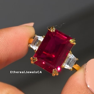 Puede incluir: Un anillo de oro con una gran gema rectangular de color rojo intenso flanqueada por dos gemas claras rectangulares más pequeñas. El anillo se sostiene sobre un fondo neutro. El texto "EtherealJewelsCA" es visible.