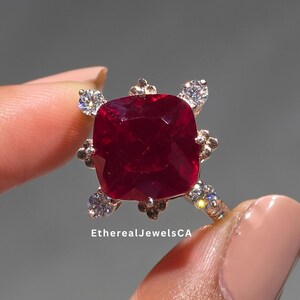 May include: A close-up of a ring featuring a large, square-cut, deep red gemstone. The ring has a gold band with small, clear accent stones and a decorative setting around the main stone. The text "EtherealJewelsCA" is visible.