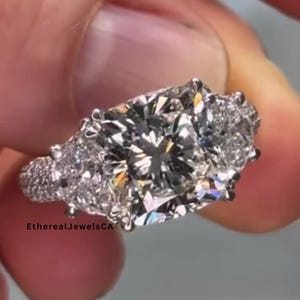 May include: A sparkling, multi-stone engagement ring with a large, square-cut center diamond and smaller accent stones. The ring features a pave band and is held up by a person's fingers. The ring is a brilliant, clear color.