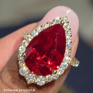 May include: A gold ring featuring a large, pear-shaped, vibrant red gemstone surrounded by a halo of smaller, clear gemstones. The ring's band is also gold. The text "EtherealJewelsCA" is visible on the image.