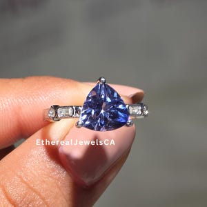 Op de afbeelding: Een zilveren ring met een grote, driehoekige, diepblauwe edelsteen als middelpunt. De ring wordt aan weerszijden geflankeerd door kleinere, rechthoekige stenen. De ring wordt door een hand vastgehouden. De tekst "EtherealJewelsCA" is zichtbaar.