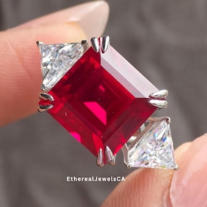 May include: A silver ring with a large, rectangular, bright red gemstone, with two smaller, white, triangular stones. The ring is held between fingers, highlighting the design and colour contrast. The text "EtherealJewelsCA" is visible.