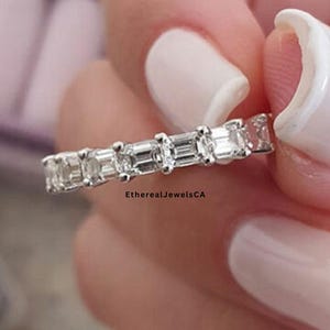 May include: A silver ring set with multiple emerald-cut clear stones. The ring is held between fingers with white nail polish. The text "EtherealJewelsCA" is visible on the ring.