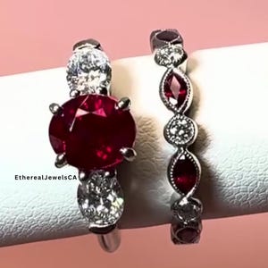 May include: Two rings with a ruby and diamond design. The first ring has a large oval ruby center stone, flanked by two smaller diamonds. The second ring features a series of ruby and diamond accents. The rings are set on a white surface against a pink background.