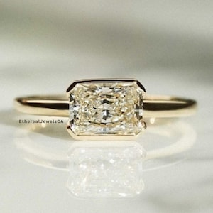 May include: A gold ring with a rectangular, light-colored gemstone. The gemstone is set in a bezel setting. The ring is on a reflective surface. The text "EtherealJewelsCA" is visible on the ring.