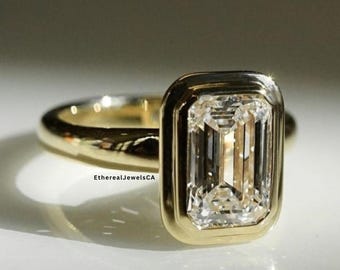 May include: A gold ring with a large, rectangular, clear gemstone. The gemstone is set in a gold bezel. The ring is on a white surface. The ring is a piece of jewelry.