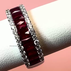 May include: A close-up of a ring with a row of rectangular, deep red gemstones set between two rows of smaller, clear stones. The ring is on a white surface with a pink background.