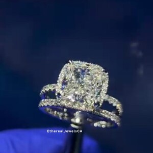 May include: A close-up of a diamond engagement ring with a large, square-shaped center stone surrounded by smaller diamonds. The ring is set in white gold and has a delicate band. The ring is held by a gloved hand against a blue background. The text "EtherealJewelsCA" is visible below the ring.