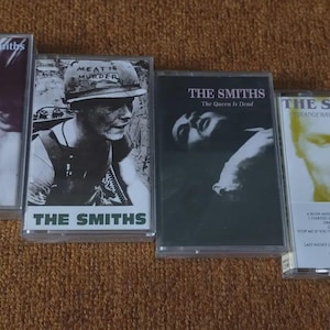May include: Four The Smiths cassette tapes. Album covers feature black and white imagery, including a person, a soldier with "MEAT IS MURDER", and a portrait. The band's name is printed on each cassette, a classic music format.