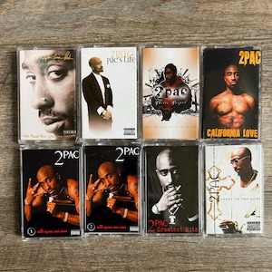 May include: A collection of eight 2Pac music cassette tapes in clear plastic cases. The covers feature various portraits of the artist, including titles like Pac's Life, Ghetto Gospel, California Love, All Eyez On Me, Greatest Hits, and Loyal To The Game.