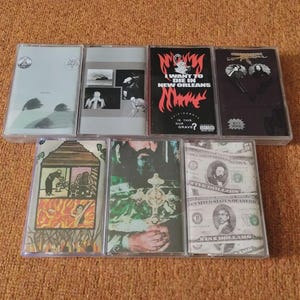 Könnte beinhalten: Sieben Musikkassetten in transparenten Hüllen. Die Kassetten zeigen verschiedene Albumcover, darunter der Text "I Want to Die in New Orleans" und Bilder von Dollarnoten. Die Kassetten liegen auf einer orangefarbenen Oberfläche.