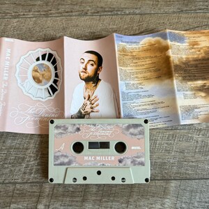 May include: A Mac Miller The Divine Feminine cassette tape with a light green shell and a pink label featuring clouds. The unfolded J-card insert displays a portrait of Mac Miller, a circular floral graphic, and printed tracklist information.