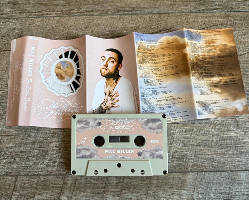 May include: A Mac Miller The Divine Feminine cassette tape with a light green shell and a pink label featuring clouds. The unfolded J-card insert displays a portrait of Mac Miller, a circular floral graphic, and printed tracklist information.