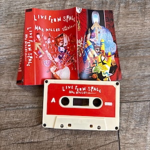 May include: A red and white cassette tape titled Live From Space by Mac Miller with The Internet, featuring a matching red j-card insert with colorful graphic illustrations of a person, a hand, and various abstract shapes and text.