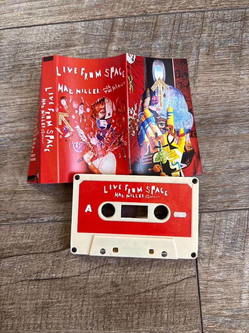 May include: A red and white cassette tape titled Live From Space by Mac Miller with The Internet, featuring a matching red j-card insert with colorful graphic illustrations of a person, a hand, and various abstract shapes and text.