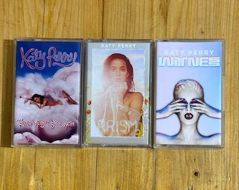 May include: Three Katy Perry cassette tapes. The first features a woman lying on a cloud with the text "Katy Perry". The second has a woman with the word "Prism". The third has a woman with the word "Witness".