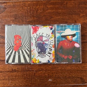 May include: Three cassette tapes featuring album art for Cage the Elephant. The left tape shows a red face on a black and white striped background with the text Melophobia. The center tape features a skull illustration. The right tape shows a person in a white hat.