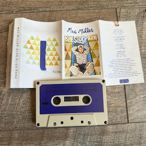 May include: A Mac Miller Blue Slide Park cassette tape with a beige shell and purple label. The unfolded j-card features a graphic of a person on a blue slide, yellow and green triangles, track listings, and production credits in black text.