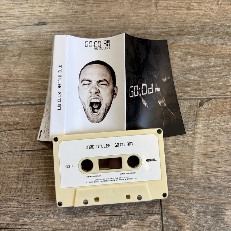 May include: A beige cassette tape labeled Mac Miller GO:OD AM Side A, accompanied by a folded paper insert featuring a black and white portrait of a person with an open mouth and the text GO:OD AM Mac Miller printed in a digital clock style font.