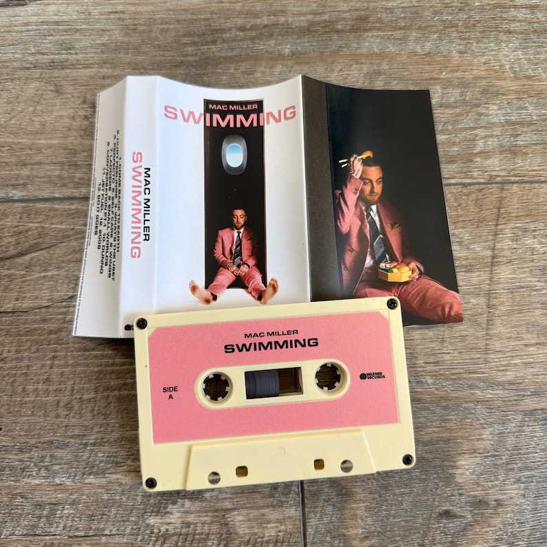 May include: A Mac Miller Swimming cassette tape with a cream-colored shell and a pink label featuring black text. The accompanying J-card insert displays the album title, tracklist, and a photograph of a person in a pink suit eating with chopsticks.