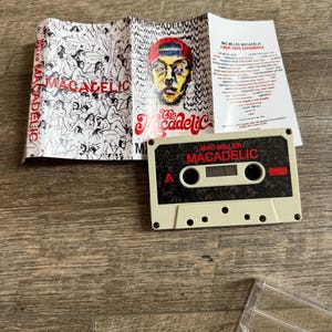 May include: A Mac Miller Macadelic cassette tape with a beige shell and black label featuring red text. The accompanying J-card insert displays a colorful portrait of a person in a red cap, a black and white illustration of figures, and a tracklist.