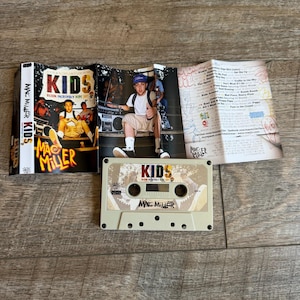 May include: A beige cassette tape featuring the Mac Miller K.I.D.S. album artwork, accompanied by a folded paper insert. The insert displays the album title, tracklist, and a photograph of the artist wearing a white t-shirt and blue cap.