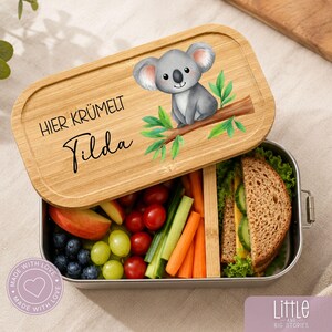 Könnte beinhalten: Eine rechteckige Brotdose aus Metall mit einem Bambusdeckel, der eine Aquarellillustration eines Koalas auf einem Ast, den Text "HIER KRÜMELT" und den Namen "Tilda" zeigt. Die Dose enthält Apfelscheiben, grüne Weintrauben, Kirschtomaten, Karottensticks und ein Sandwich.